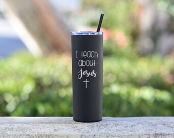 May include: A black stainless steel tumbler with a white inscription that reads "I teach about Jesus" and a small cross below the text. The tumbler has a black lid and a black straw.