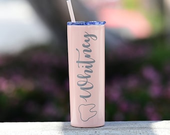 May include: A pink stainless steel tumbler with a blue lid and a white straw. The tumbler has a gray tooth graphic and the name "Whitney" written in cursive.