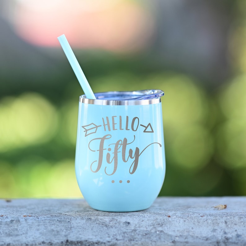 Puede incluir: Un vaso de vino de acero inoxidable azul claro con tapa plateada y pajita. El vaso tiene el texto "Hello Fifty" impreso en &eacute;l.