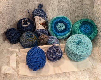 May include: A collection of blue and teal yarn balls, some with a multi-colored pattern, and a skein of yarn with the label "Impeccable" on a white surface.