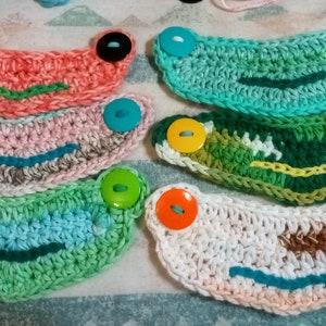 Puede incluir: Un conjunto de ocho extensores de mascarilla de crochet con botones coloridos. Los extensores están hechos en una variedad de colores, incluyendo rosa, azul, verde y blanco. Los botones también son coloridos, algunos son amarillos, naranjas y verdes.