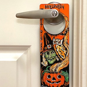 May include: Halloween door hanger with a witch holding a jack-o-lantern. The witch is wearing a black hat and green dress. An owl is perched on her shoulder. The text "Halloween" is at the top of the hanger.