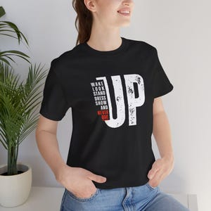 May include: Black t-shirt with the word "UP" in large white distressed font. Below the "UP" are the words "WAKE LOOK STAND DRESS SHOW AND NEVER GIVE" in white and red font. The shirt is made of a soft material.