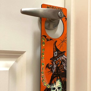 May include: Orange and black Halloween door hanger with a witch holding a skull. The text on the hanger reads "Halloween Do Not Disturb".