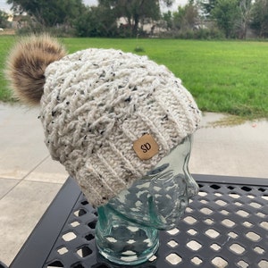 May include: White crocheted beanie with a brown faux fur pom pom and a brown leather tag with the letters "SO" on it.