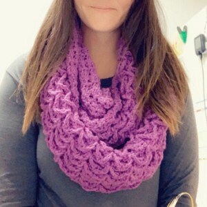 May include: A handmade, lavender-colored crochet infinity scarf. The scarf is made with a textured stitch, creating a woven appearance. The scarf is worn around the neck and is a solid color. The scarf is a cozy accessory.