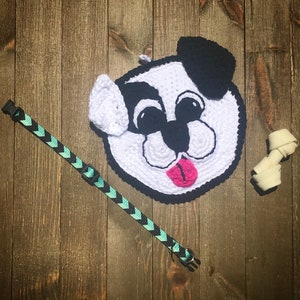 May include: A white crocheted dog with black ears and a pink tongue. The dog is lying on a wooden surface with a blue and green patterned dog collar and a white bone.