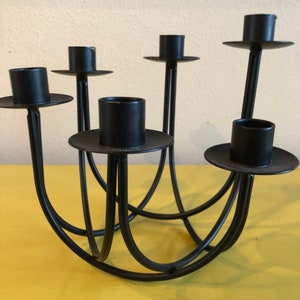 Puede incluir: Un candelabro de metal negro con seis portavelas. Los soportes son circulares y se asientan sobre brazos delgados y curvos. La base es una serie de arcos interconectados. El candelabro est&aacute; dise&ntilde;ado para sostener m&uacute;ltiples velas.