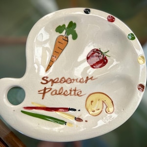 May include: A white ceramic spoon rest in the shape of a painter's palette. It features hand-painted illustrations of a carrot, tomato, and a fortune cookie. The words "Spooner Palette" are written in brown script.