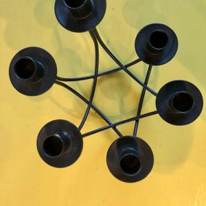 Puede incluir: Candelabro de metal negro con cinco soportes circulares dispuestos en forma de estrella. Los portavelas est&aacute;n conectados por l&iacute;neas met&aacute;licas curvas. El fondo es de color amarillo liso.