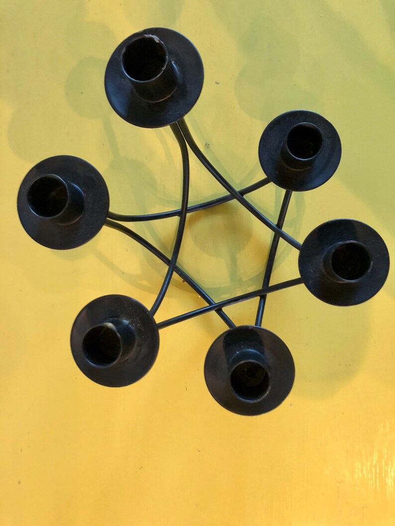 Puede incluir: Candelabro de metal negro con cinco soportes circulares dispuestos en forma de estrella. Los portavelas est&aacute;n conectados por l&iacute;neas met&aacute;licas curvas. El fondo es de color amarillo liso.