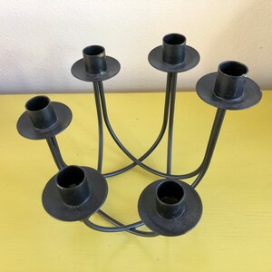 Puede incluir: Un candelabro de metal negro con cinco portavelas. Los soportes son circulares y se asientan sobre brazos curvos y delgados que se cruzan en el centro. El candelabro est&aacute; sobre una superficie amarilla.