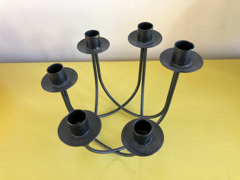 Puede incluir: Un candelabro de metal negro con cinco portavelas. Los soportes son circulares y se asientan sobre brazos curvos y delgados que se cruzan en el centro. El candelabro est&aacute; sobre una superficie amarilla.