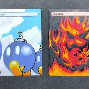 May include: Two hand-painted cards. One features blue bombs with yellow accents against a blue sky, labeled "Island." The other depicts a fiery, red and orange design on a black background, labeled "Mountain."