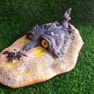 Può includere: Un diorama in miniatura che raffigura una creatura testurizzata grigio scuro con due grandi occhi rotondi e chiari che emerge da una base color sabbia. Un piccolo cavallo scuro con un cavaliere è posizionato sulla sabbia vicino alla creatura.