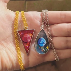 Puede incluir: Dos collares con colgantes de resina. Uno es un triángulo rojo con un diseño de gema facetada y copos dorados en una cadena dorada. El otro tiene forma de lágrima con un diseño de gema azul y purpurina en una cadena plateada con marco metálico.