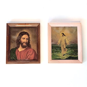 May include: Two framed religious prints. One features a portrait of a man with long hair and a beard, wearing a red robe and green shawl, in a brown wooden frame. The other depicts a figure walking on water, in a pink frame.