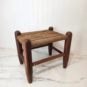 May include: A small footstool featuring a dark brown wooden frame with four rounded legs and a rectangular seat woven from natural tan fibers. The stool has a sturdy construction with horizontal support bars connecting the legs on all sides.