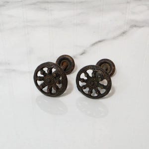 May include: Two circular metal drawer knobs with a dark, weathered bronze finish. Each knob features an openwork design with a central floral motif surrounded by eight radiating spokes and a textured outer rim, attached to a short threaded post.