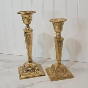 May include: Two polished brass candlesticks of varying heights, each featuring a square base, a tapered stem with embossed floral and ribbon motifs, and a wide, flared candle cup with decorative detailing around the upper rim.