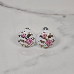 May include: Two round white ceramic cabinet knobs featuring a floral pattern with pink rose blossoms and metallic gold leaves. Each knob has a silver-toned metal post extending from the back, designed for attachment to furniture or cabinetry doors.