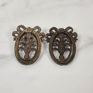 May include: Two oval-shaped brass drawer pulls featuring a decorative ribbon bow at the top and an intricate openwork floral design in the center. The metal has a darkened, aged patina with visible highlights on the raised edges of the hardware.