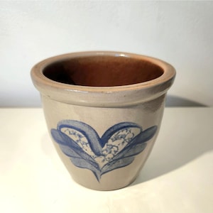 May include: A stoneware planter with a light beige exterior and a brown interior. The planter features a blue floral design on the front. The rim is rounded and the planter has a slightly tapered shape. This decorative planter is suitable for indoor or outdoor use.