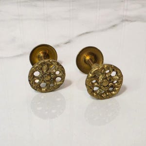 May include: Two circular brass cabinet knobs featuring a textured, openwork floral pattern. Each knob has a central stem and a flat circular base, showcasing a weathered, metallic gold finish with intricate cutouts across the face.
