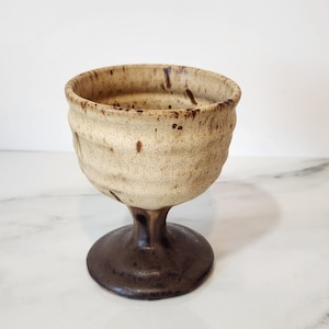May include: A handmade ceramic goblet with a textured, off-white bowl and a dark brown stem and base. The goblet has a rustic, organic appearance, with visible speckles and variations in color. The design is simple and elegant.