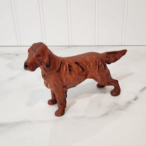 May include: A brown ceramic dog figurine in a standing pose. The dog has a long snout, floppy ears, and a curled tail. The figurine is painted with dark brown accents and has a glossy finish. The dog is a reddish-brown color.