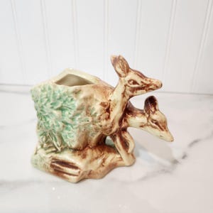 May include: A ceramic planter featuring two deer figures. The planter is tan and brown with green foliage accents. The deer are light brown with detailed features. The planter has a rectangular opening for plants.