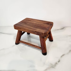 May include: A small, handcrafted wooden stool with a square top and four angled legs. The stool is made of dark brown wood with a natural grain pattern. The legs have black rubber feet. This decorative stool is perfect for home decor.