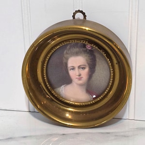 May include: A circular brass frame containing a miniature portrait of a person with styled hair, a small pink flower accessory, and a light-colored garment. The frame features a decorative inner border and a small scalloped hanging loop at the top.