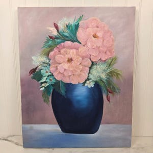 May include: An oil painting on canvas depicting a bouquet of large, layered pink flowers and small white blossoms with dark green foliage arranged in a smooth, deep navy blue vase. The background is a soft, muted mauve with a light blue surface below.