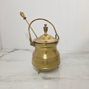 May include: A polished brass pot with a rounded body, three small feet, and a matching lid featuring a central finial. A curved handle is attached to the sides, and a long, slender brass spoon with a decorative top rests inside the pot.