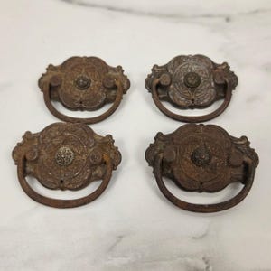 May include: Four matching metal drawer pulls with a dark, weathered brown finish. Each pull features an ornate, circular backplate with embossed floral patterns and a central rosette, paired with a simple, rounded, hinged drop handle.