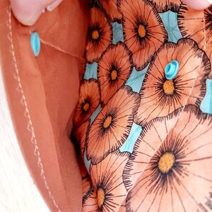 May include: Close-up of a peach-colored fabric pouch with a floral pattern. The interior features a light blue background with large orange and black flower illustrations. The pouch is secured with a light blue snap closure.