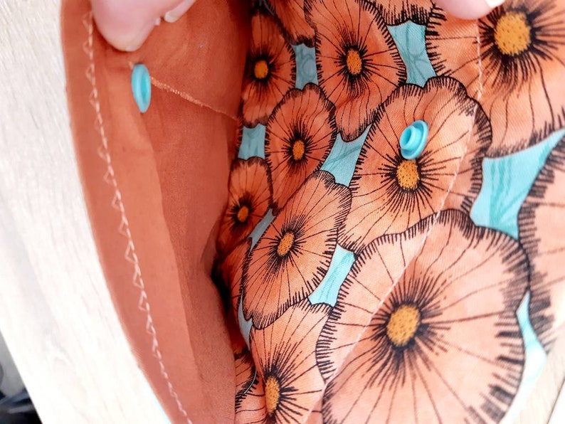 May include: Close-up of a peach-colored fabric pouch with a floral pattern. The interior features a light blue background with large orange and black flower illustrations. The pouch is secured with a light blue snap closure.