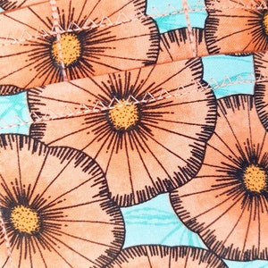 May include: Close-up of a fabric with a repeating floral pattern. The flowers are peach with dark brown outlines and yellow centers. The background is a light turquoise color. The fabric is stitched together.