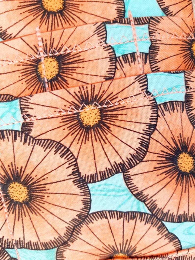 May include: Close-up of a fabric with a repeating floral pattern. The flowers are peach with dark brown outlines and yellow centers. The background is a light turquoise color. The fabric is stitched together.