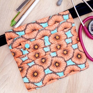 May include: A floral print medical accessory bag with a light blue background and orange flower design. A pink stethoscope and pens are next to the bag. The bag is approximately 8 inches wide.