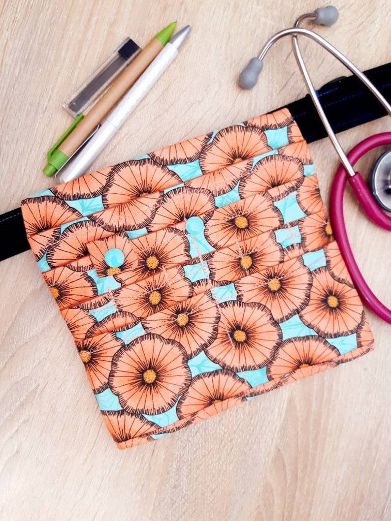 May include: A floral print medical accessory bag with a light blue background and orange flower design. A pink stethoscope and pens are next to the bag. The bag is approximately 8 inches wide.