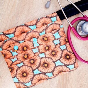 May include: A patterned medical accessory pouch with a button closure, featuring a floral design in peach and brown on a light blue background. A pink stethoscope rests beside the pouch on a wooden surface.