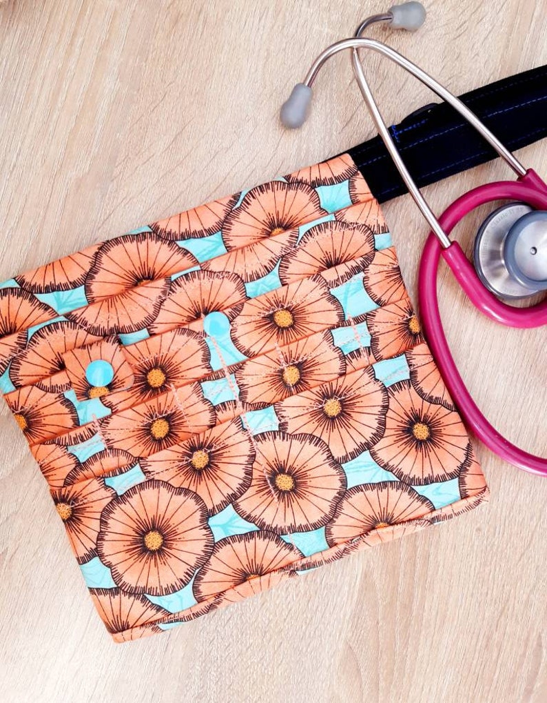 May include: A patterned medical accessory pouch with a button closure, featuring a floral design in peach and brown on a light blue background. A pink stethoscope rests beside the pouch on a wooden surface.