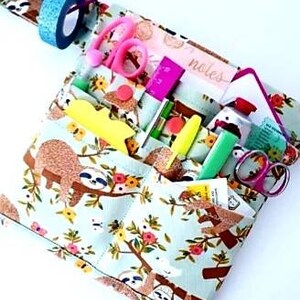 May include: A light blue fabric organizer with a sloth and floral pattern, filled with stationery items. Includes pink scissors, a pink ruler, yellow highlighters, and a roll of blue glitter tape. The word "notes" is visible.