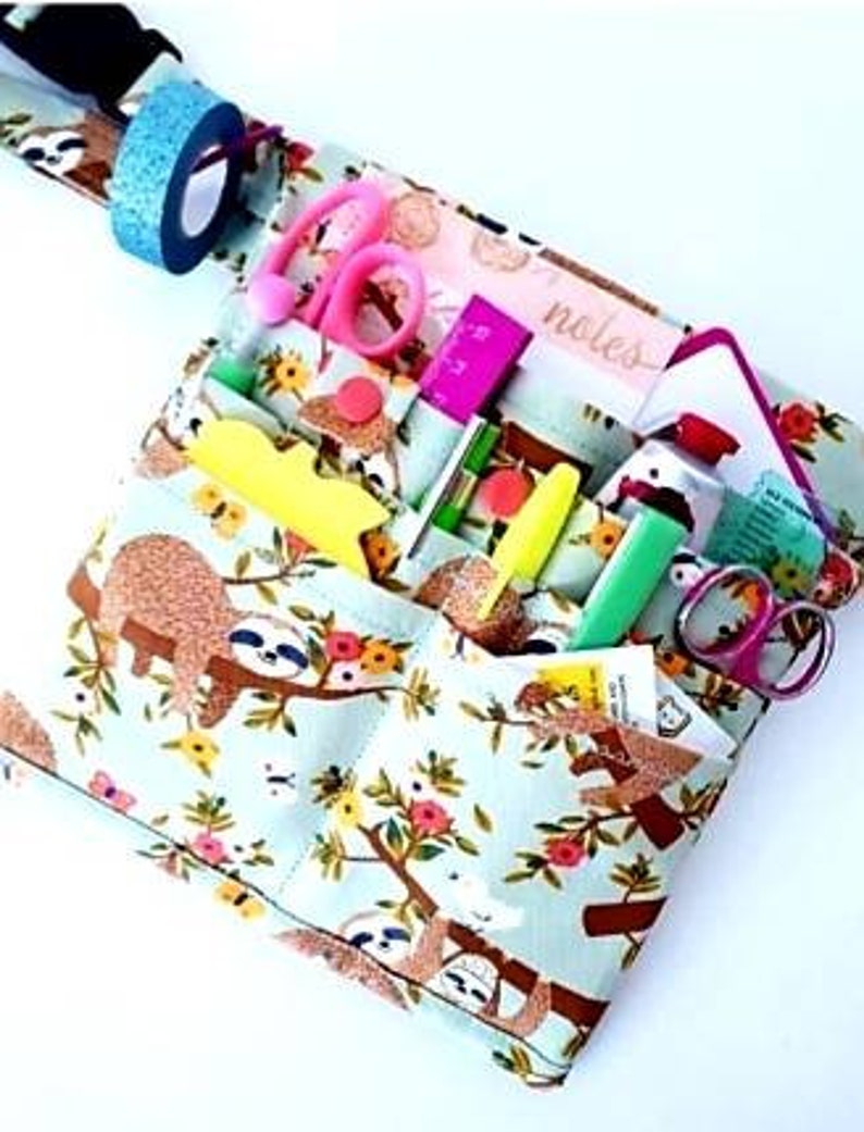 May include: A light blue fabric organizer with a sloth and floral pattern, filled with stationery items. Includes pink scissors, a pink ruler, yellow highlighters, and a roll of blue glitter tape. The word "notes" is visible.