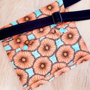 May include: A floral print fanny pack with a navy blue adjustable strap and buckle. The bag features a repeating pattern of orange flowers on a light blue background. The strap is stitched with blue thread.