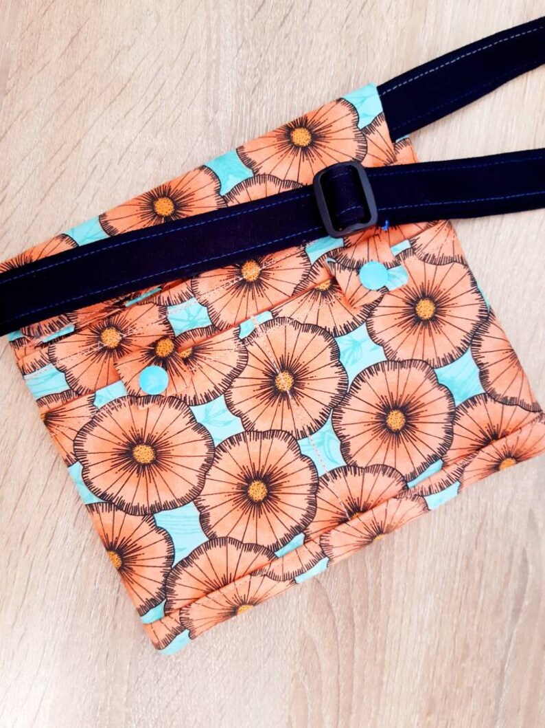 May include: A floral print fanny pack with a navy blue adjustable strap and buckle. The bag features a repeating pattern of orange flowers on a light blue background. The strap is stitched with blue thread.