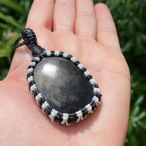 K&ouml;nnte beinhalten: Ein Anh&auml;nger aus schwarzem Obsidian mit einer Makramee-Kordel. Der ovale Stein ist in einem schwarz-wei&szlig;en Webmuster gefasst. Der Anh&auml;nger wird in einer Hand vor einem verschwommenen gr&uuml;nen Hintergrund gehalten.