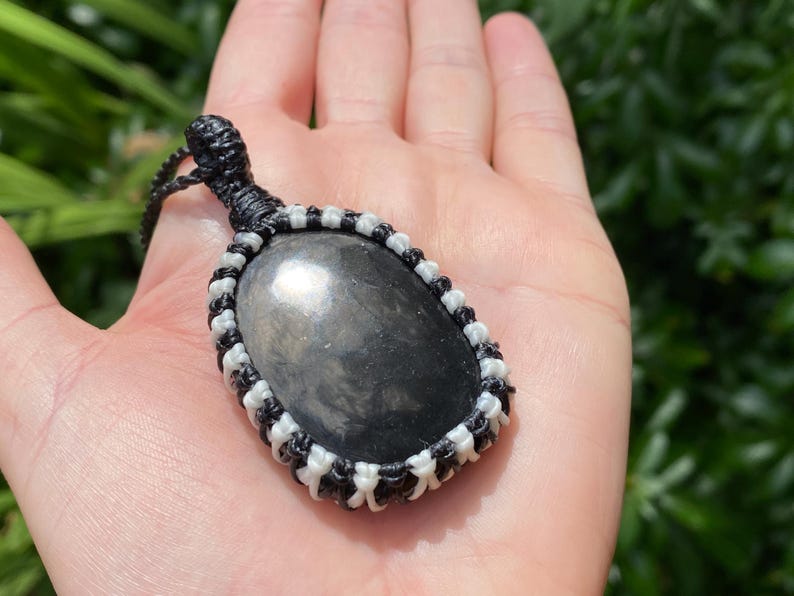 K&ouml;nnte beinhalten: Ein Anh&auml;nger aus schwarzem Obsidian mit einer Makramee-Kordel. Der ovale Stein ist in einem schwarz-wei&szlig;en Webmuster gefasst. Der Anh&auml;nger wird in einer Hand vor einem verschwommenen gr&uuml;nen Hintergrund gehalten.